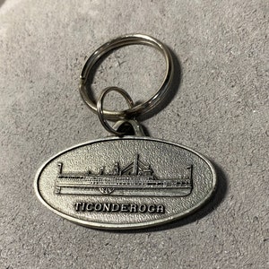 Puede incluir: Un llavero de plata con un anillo redondo y un dije ovalado. El dije presenta una imagen en blanco y negro de un barco con la palabra "TICONDEROGA" impresa debajo.