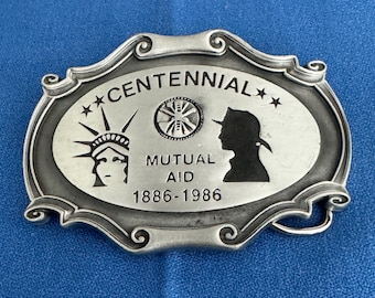 May include: Silver belt buckle with a raised design featuring a silhouette of the Statue of Liberty, a wheel, and a woman's profile. The buckle also includes the text "Centennial", "Mutual Aid", and "1886-1986".