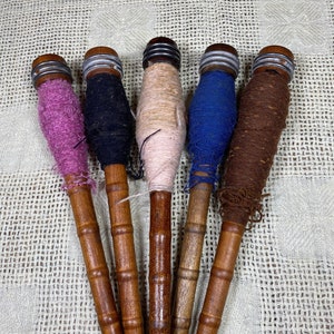 Può includere: Cinque bobine di legno con fili di diversi colori avvolti attorno. Le bobine sono in legno marrone e hanno tappi metallici alle estremità. I fili sono rosa, nero, beige, blu e marrone.