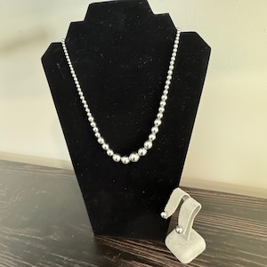 May include: A silver necklace with round beads and matching silver earrings. The necklace is displayed on a black velvet stand and the earrings are on a gray stand.