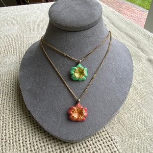 Puede incluir: Dos collares de cadena dorada con colgantes de flores. El colgante superior es verde con un centro amarillo. El colgante inferior es naranja con un centro amarillo.