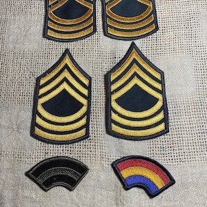 May include: Five embroidered patches, including four military rank patches with gold and black chevron designs and one black patch with a curved shape. There is also a colorful patch with red, yellow, and blue stripes.