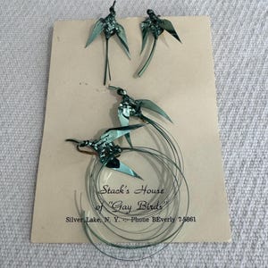 May include: Vintage metal bird ornaments in shades of green are displayed on a tan card. The card features the text "Stack's House of 'Gay Birds'" and a phone number. The birds have outstretched wings and long tails.