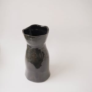 Könnte beinhalten: Eine schwarze Keramikvase mit einem unregelmäßigen, wellenförmigen Rand und einem leicht ausgestellten Boden.