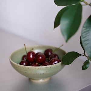 Könnte beinhalten: Eine hellgrüne Keramikschale gefüllt mit reifen, dunkelroten Kirschen. Die Schale steht auf einer grauen Oberfläche, mit grünen Blättern im Hintergrund. Die Kirschen haben lange grüne Stiele.