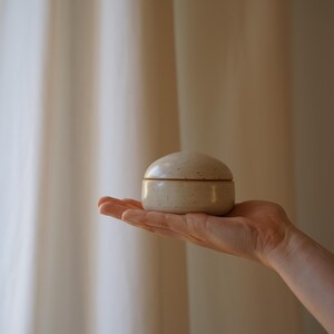 Könnte beinhalten: Kleine, gesprenkelte Keramik-Schmuckdose mit rundem Deckel. Die Dose hat eine hellbeige Farbe und wird in der Hand einer Person gehalten. Der Hintergrund ist ein neutral getönter Vorhang.