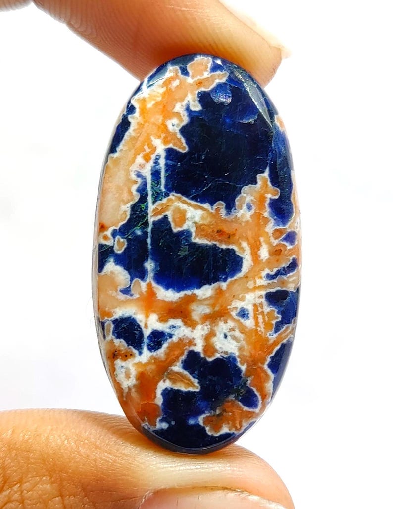 May include: An oval-shaped cabochon gemstone featuring a deep blue base colour with intricate, branching patterns in shades of orange and white. The stone has a smooth, polished surface and a distinct, natural-looking vein structure throughout.
