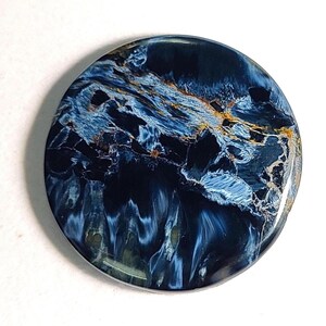 Puede incluir: Un cabuj&oacute;n redondo de ojo de tigre azul y dorado. La piedra tiene un patr&oacute;n de remolino de azul, dorado y negro.