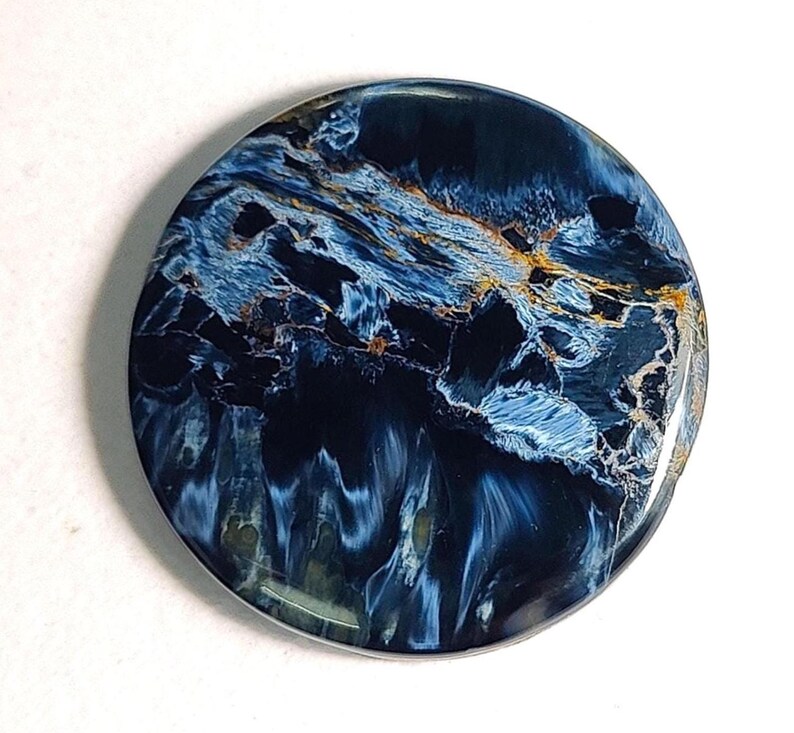 Puede incluir: Un cabuj&oacute;n redondo de ojo de tigre azul y dorado. La piedra tiene un patr&oacute;n de remolino de azul, dorado y negro.