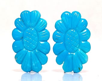 May include: Two turquoise-colored, carved flower-shaped gemstones. Each gemstone features a central oval with etched lines and surrounding petals. The gemstones are a bright, solid blue color and are set against a white background.
