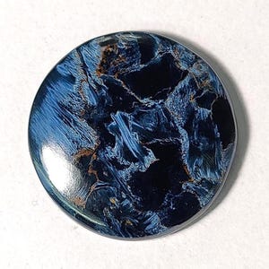 Puede incluir: Un cabuj&oacute;n redondo de ojo de tigre azul y dorado. La piedra tiene un patr&oacute;n de remolino de azul y dorado.