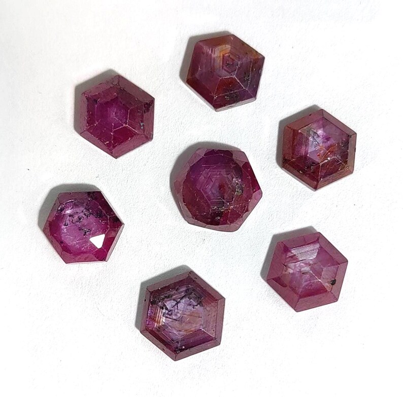 May include: Seven faceted, hexagonal-shaped, deep red gemstones. The gems have a polished appearance with visible internal structures and variations in colour. The arrangement is on a white surface, showcasing the gems' rich colour and cut.
