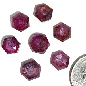May include: Seven faceted, hexagonal, deep red ruby gemstones are displayed on a white surface. The gemstones have a polished appearance and varying internal patterns. A coin is included for size comparison.