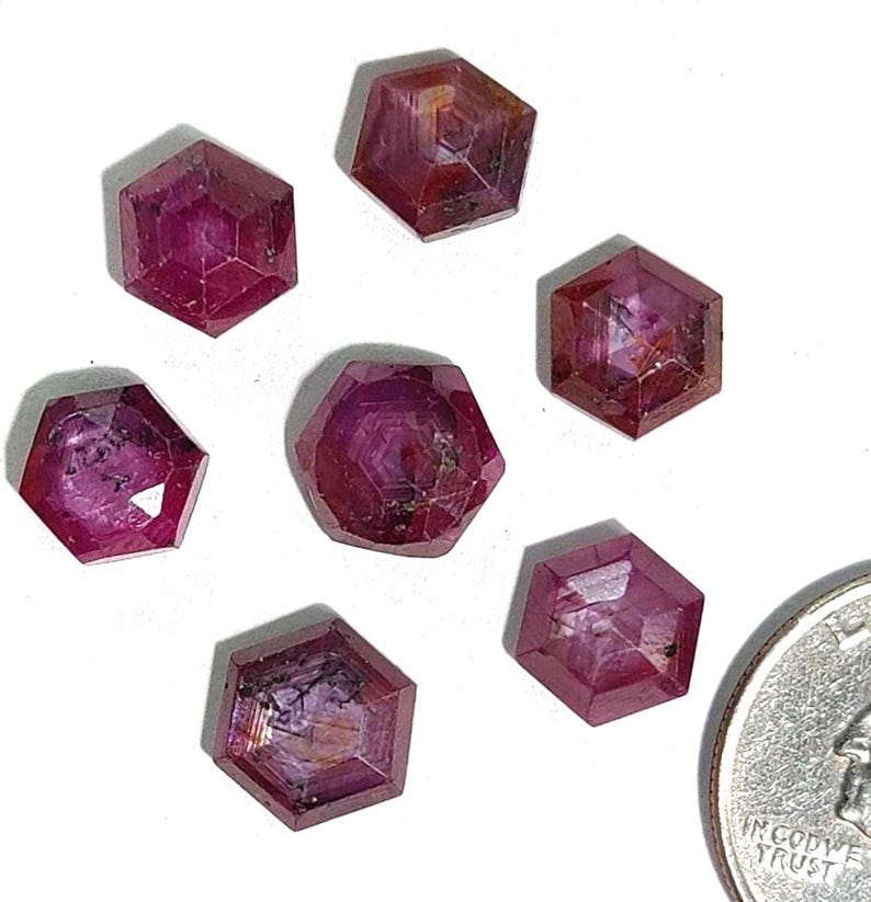 May include: Seven faceted, hexagonal, deep red ruby gemstones are displayed on a white surface. The gemstones have a polished appearance and varying internal patterns. A coin is included for size comparison.