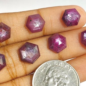 May include: Seven hexagonal, faceted ruby gemstones in shades of red and pink are displayed on a hand. A UK coin is next to the gems for size comparison. The gemstones have a polished, reflective surface and are of varying sizes.