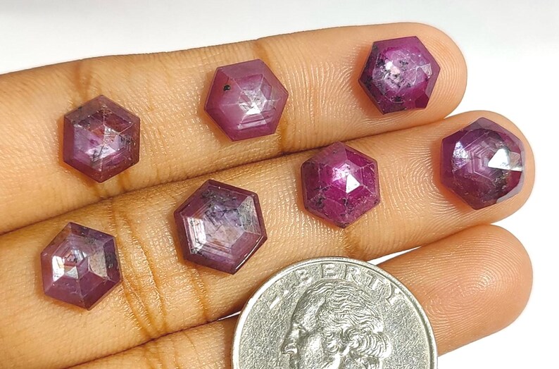 May include: Seven hexagonal, faceted ruby gemstones in shades of red and pink are displayed on a hand. A UK coin is next to the gems for size comparison. The gemstones have a polished, reflective surface and are of varying sizes.