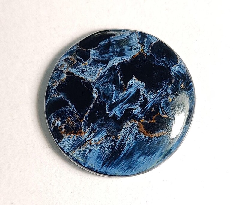 Puede incluir: Un cabuj&oacute;n redondo de ojo de tigre azul y dorado. La piedra tiene un patr&oacute;n de remolino de azul, negro y dorado.
