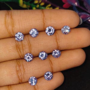 May include: A collection of ten round, light blue gemstones with multiple facets, displayed on a hand. The gems sparkle, reflecting light. The background is dark, emphasizing the gems' brilliance. The gems are likely for jewelry making.