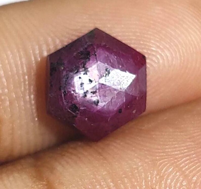 May include: A faceted, hexagonal ruby gemstone. The gem is a deep, saturated red with dark inclusions. The facets catch the light, creating a sparkling effect. The gemstone is held in a hand.