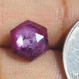 May include: A faceted, hexagonal ruby gemstone with a deep magenta hue. The gem has multiple reflective surfaces and dark inclusions. The ruby is next to a silver coin.