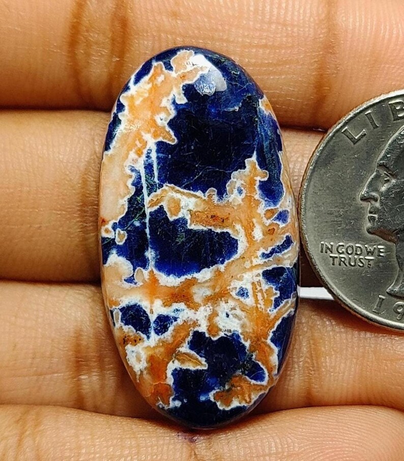 May include: An oval-shaped cabochon gemstone featuring a deep blue base colour with intricate, branching patterns in shades of light orange and white. The stone is positioned next to a United States quarter coin for scale, highlighting its size.