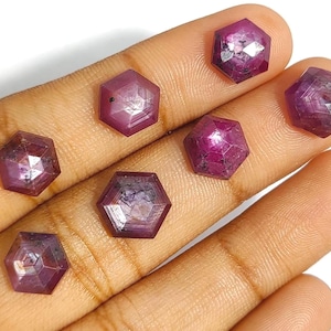 Puede incluir: Siete gemas facetadas, de forma hexagonal, de color rojo intenso. Las gemas se sostienen en una mano, y cada piedra tiene un patrón interno único. Las facetas captan la luz, creando un efecto brillante. Probablemente rubíes.