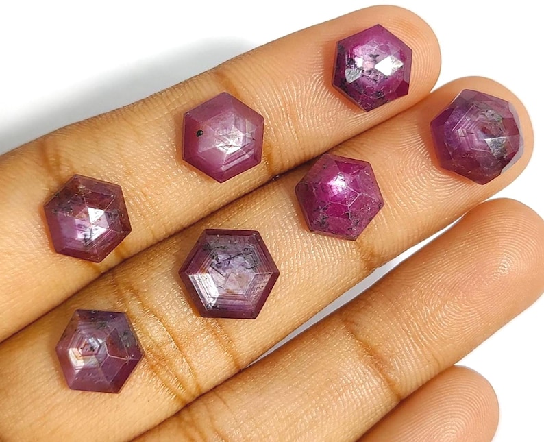 May include: Seven faceted, hexagonal-shaped, deep red gemstones. The gems are held in a hand, and each stone has a unique internal pattern. The facets catch the light, creating a sparkling effect. These gemstones are likely rubies.