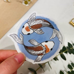 May include: A sticker featuring two koi fish in a yin-yang arrangement. The fish have white bodies with orange and black markings, set against a light blue watercolor background. The sticker is round and has a glossy finish.