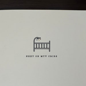 May include: A white card with a black line drawing of a baby crib and mobile. The text "NEXT ON MTV CRIBS" is printed below the crib in a simple sans-serif font. The card is on a brown surface.
