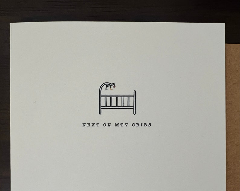 May include: A white card with a black line drawing of a baby crib and mobile. The text "NEXT ON MTV CRIBS" is printed below the crib in a simple sans-serif font. The card is on a brown surface.