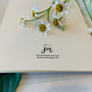 May include: A cream-colored greeting card with the text "LOVE BY jm" and the website address. The card is adorned with fresh daisies with white petals and yellow centers. The words "Made in Brooklyn, NY" are also printed on the card.