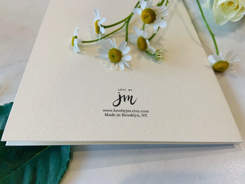 May include: A cream-colored greeting card with the text "LOVE BY jm" and the website address. The card is adorned with fresh daisies with white petals and yellow centers. The words "Made in Brooklyn, NY" are also printed on the card.