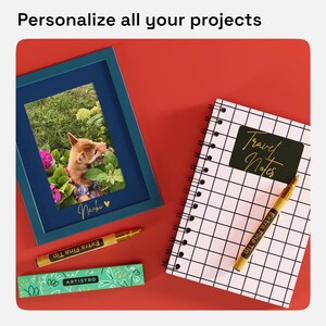 May include: A blue picture frame with a photo of a dog in a grassy area with pink flowers. The frame has the word "Naoki" written in gold lettering. A gold pen with the words "Extra Fine Tip" is lying on a green and white box with the word "Artistro" on it. A white notebook with a black grid pattern has a black sticker with the words "Travel Notes" written in gold lettering. A gold pen with the words "Extra Fine Tip" is lying on the notebook.