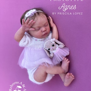 May include: A sleeping baby doll wearing a white knit outfit with a tulle skirt and a matching headband. A crocheted bunny doll with a pink tutu rests beside the baby. The text "Prototype Agnes by Priscila Lopez" is visible.
