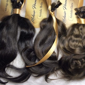 May include: Three bundles of human hair extensions in different shades of brown. The hair is tied with a gold ribbon and labeled with the brand name "Mohair Premium" and the color name in Spanish.