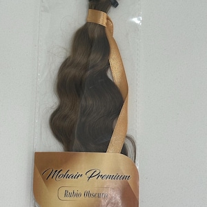 May include: A package of brown wavy human hair extensions with a gold ribbon and a label that reads "Mohair Premium Rubio Obscuro".