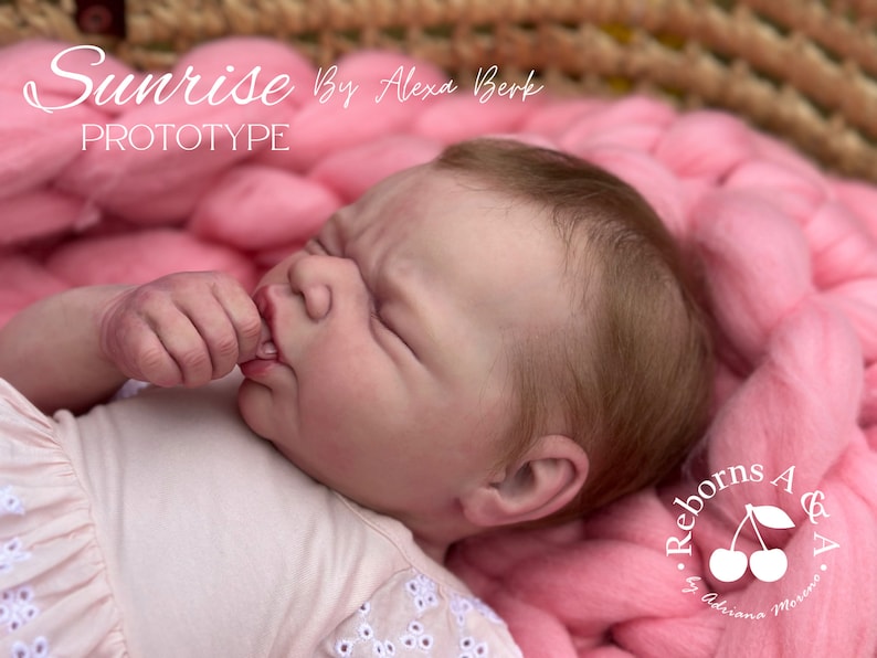 Puede incluir: Una mu&ntilde;eca beb&eacute; realista, con piel clara y cabello casta&ntilde;o claro, acostada sobre una manta rosa tejida. La mu&ntilde;eca lleva un vestido rosa claro. El texto "Sunrise Prototype" es visible en la imagen.