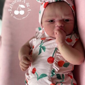 Puede incluir: Un beb&eacute; reborn con un conjunto rosa con estampado de cerezas y una diadema a juego. La mu&ntilde;eca tiene una expresi&oacute;n seria y est&aacute; acostada sobre una superficie rosa. El texto "Reborns A & A by Adriana Moreno" est&aacute; en la esquina superior izquierda.
