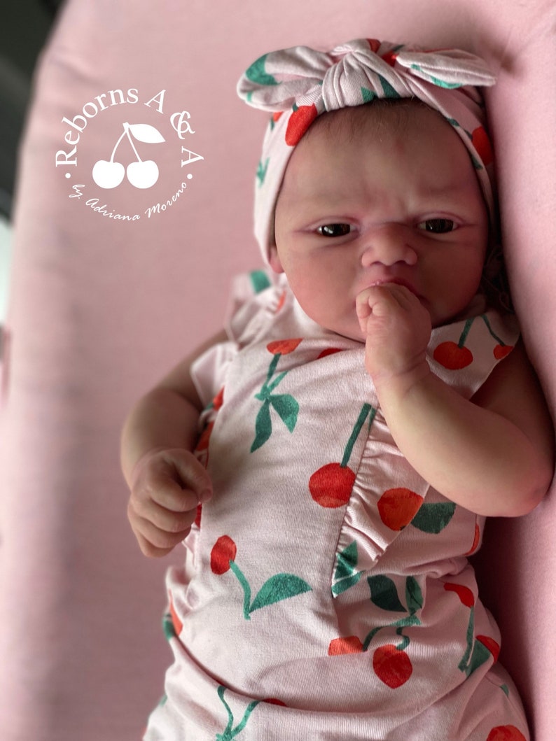 Puede incluir: Un beb&eacute; reborn con un conjunto rosa con estampado de cerezas y una diadema a juego. La mu&ntilde;eca tiene una expresi&oacute;n seria y est&aacute; acostada sobre una superficie rosa. El texto "Reborns A & A by Adriana Moreno" est&aacute; en la esquina superior izquierda.