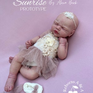Puede incluir: Una mu&ntilde;eca beb&eacute; realista, vestida con un top color crema con adornos florales y de perlas y una falda de tul rosa. La mu&ntilde;eca tiene una diadema blanca con un acento floral y est&aacute; sobre una superficie rosa. El texto "Sunrise By Alexa Berk Prototype" es visible.