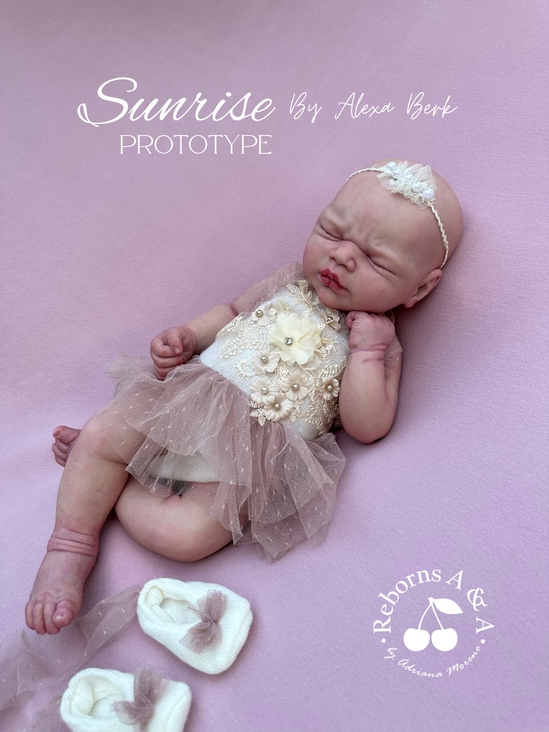 Puede incluir: Una mu&ntilde;eca beb&eacute; realista, vestida con un top color crema con adornos florales y de perlas y una falda de tul rosa. La mu&ntilde;eca tiene una diadema blanca con un acento floral y est&aacute; sobre una superficie rosa. El texto "Sunrise By Alexa Berk Prototype" es visible.