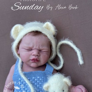 Puede incluir: Una mu&ntilde;eca reborn con un pelele de punto azul claro y un gorro de oso color crema con detalles de orejas. La mu&ntilde;eca sostiene un peque&ntilde;o osito de peluche color crema. El texto "PROTOTYPE Sunday By Alexa Berk" es visible.