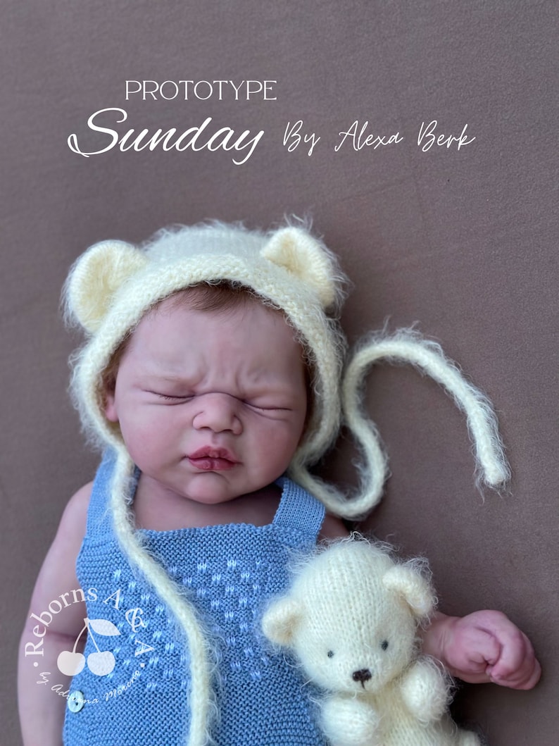 Puede incluir: Una mu&ntilde;eca reborn con un pelele de punto azul claro y un gorro de oso color crema con detalles de orejas. La mu&ntilde;eca sostiene un peque&ntilde;o osito de peluche color crema. El texto "PROTOTYPE Sunday By Alexa Berk" es visible.
