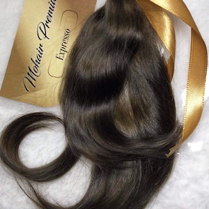 Op de afbeelding: Een bundel donkerbruine hairextensions, vastgebonden met een gouden lint. Een gouden label is bevestigd, met de woorden "Mohair Premium Expresso" erop gedrukt. Het haar heeft een natuurlijke golf en glans.