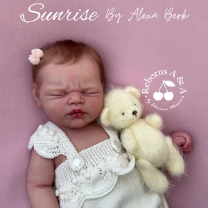 Puede incluir: Una mu&ntilde;eca reborn llamada "Sunrise" de Alexa Berk, con un pelele blanco tejido y un peque&ntilde;o osito de peluche color crema. La mu&ntilde;eca tiene los ojos cerrados, un accesorio para el pelo de flor rosa, sobre fondo rosa.