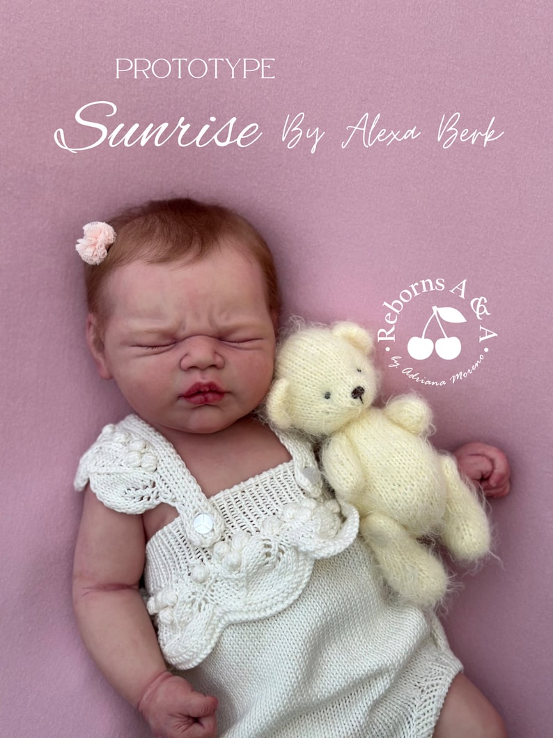 Puede incluir: Una mu&ntilde;eca reborn llamada "Sunrise" de Alexa Berk, con un pelele blanco tejido y un peque&ntilde;o osito de peluche color crema. La mu&ntilde;eca tiene los ojos cerrados, un accesorio para el pelo de flor rosa, sobre fondo rosa.