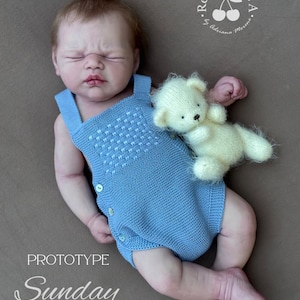 Puede incluir: Una mu&ntilde;eca beb&eacute; con un pelele de punto azul claro con detalles de botones, tumbada sobre una superficie marr&oacute;n. Un peque&ntilde;o osito de peluche color crema descansa junto a la mu&ntilde;eca. El texto "Prototype Sunday By Alexa Berk" es visible.