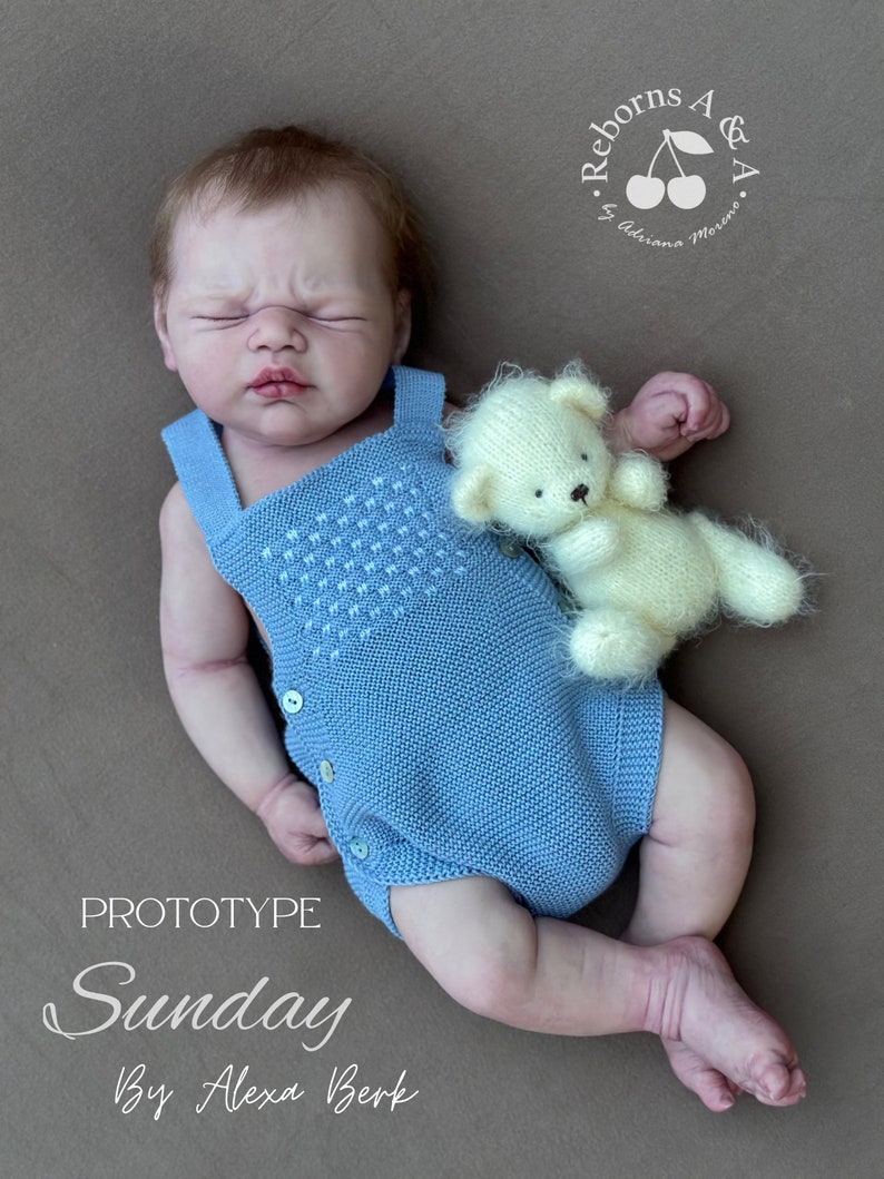 Puede incluir: Una mu&ntilde;eca beb&eacute; con un pelele de punto azul claro con detalles de botones, tumbada sobre una superficie marr&oacute;n. Un peque&ntilde;o osito de peluche color crema descansa junto a la mu&ntilde;eca. El texto "Prototype Sunday By Alexa Berk" es visible.