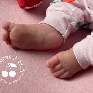 Puede incluir: Primer plano de un pie y una pierna de beb&eacute; con pijama rosa con estampado de cerezas, sobre una superficie rosa. El texto "Reborns A & A by Adriana Moreno" es visible.