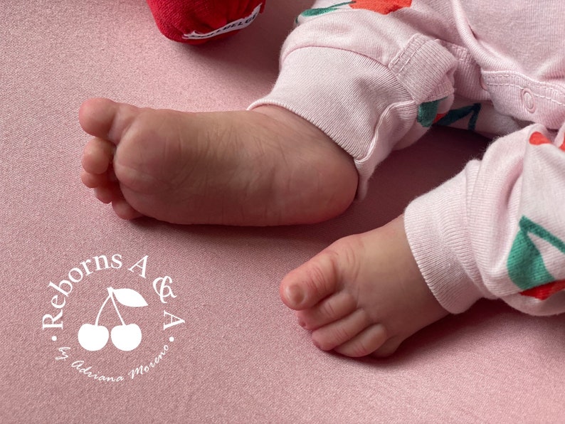 Puede incluir: Primer plano de un pie y una pierna de beb&eacute; con pijama rosa con estampado de cerezas, sobre una superficie rosa. El texto "Reborns A & A by Adriana Moreno" es visible.